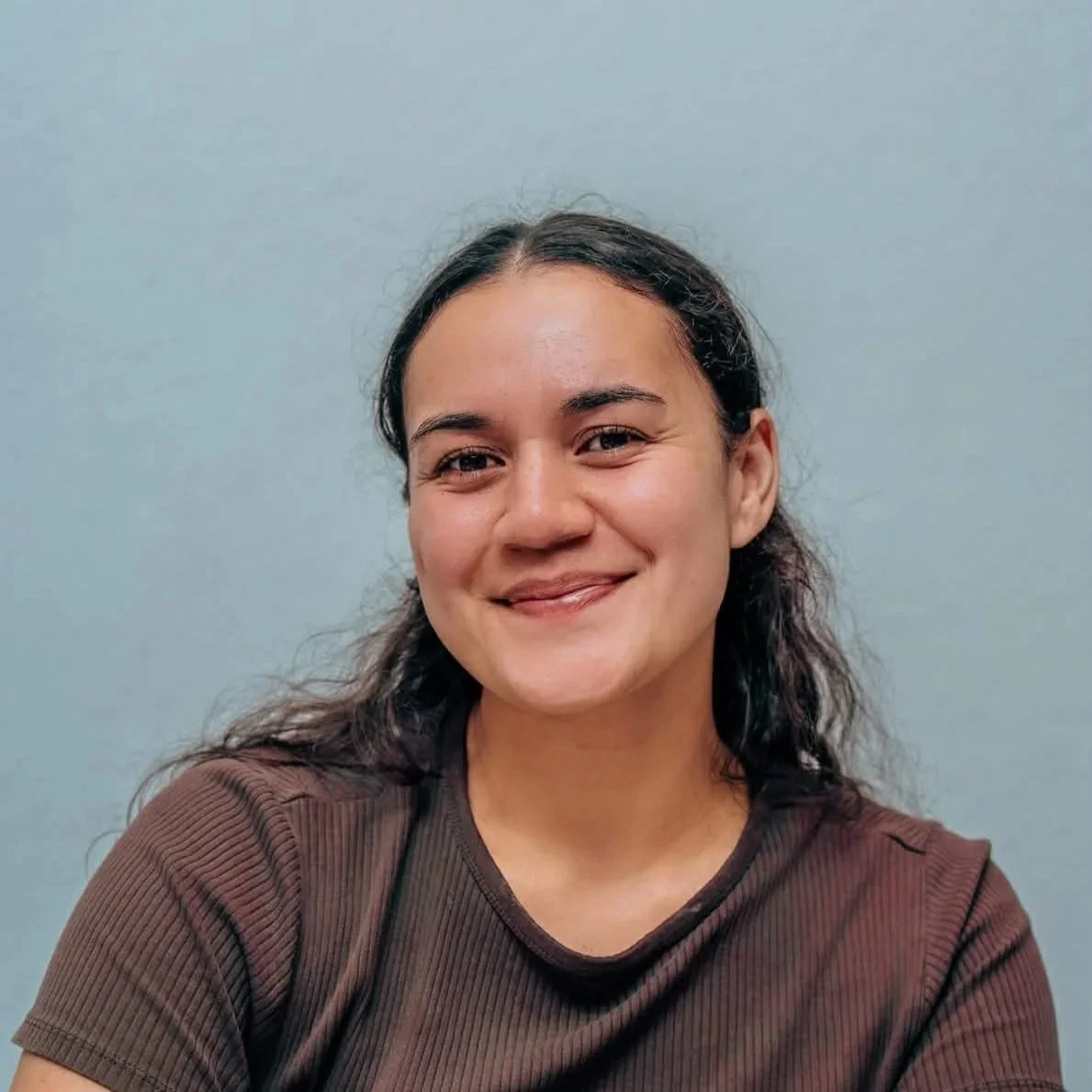 Portrait of a young woman with dark, curly hair, smiling, wearing a brown top, against a plain light blue background.