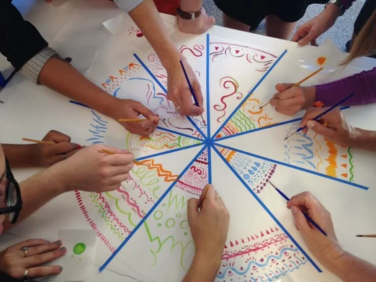 Several people are drawing and coloring on a large circular paper with various colorful patterns using paintbrushes and markers.