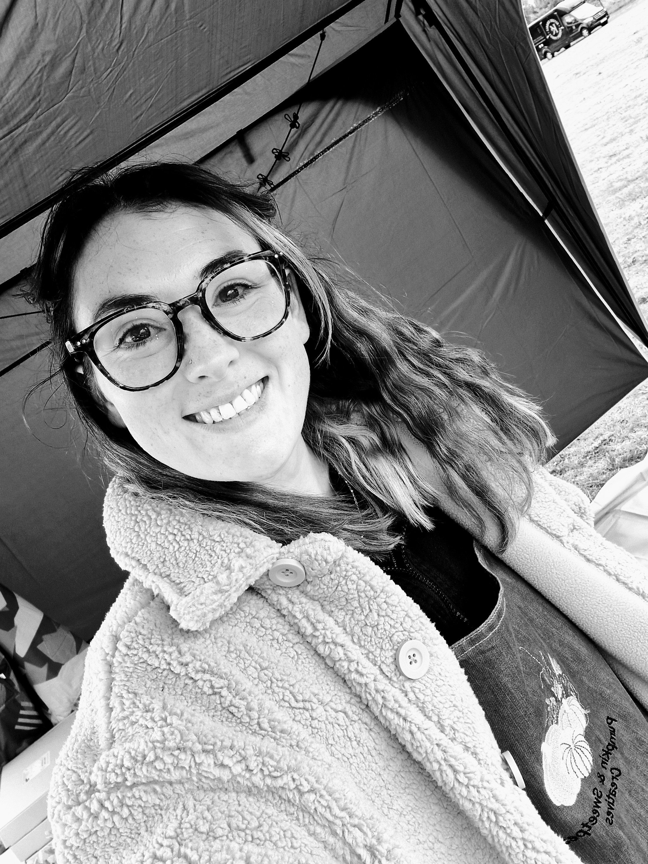 A smiling woman with glasses and wavy hair, wearing a cozy, fluffy coat, standing outdoors near a tent. The background shows some vehicles and a cloudy sky.