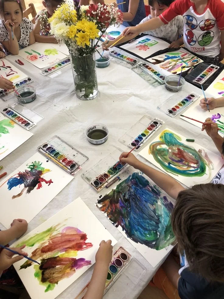 Children painting colorful pictures around a vase of flowers on a table.