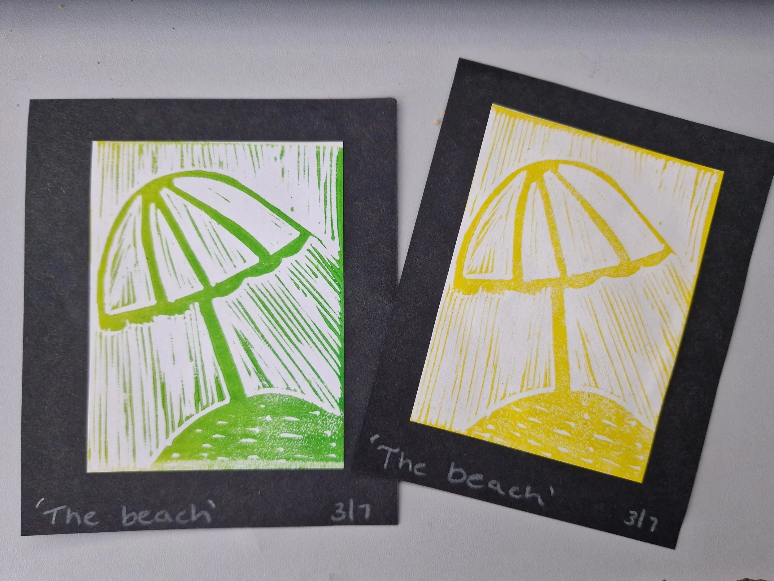 Two printed images of a beach umbrella, one in green and one in yellow, on black background paper with handwritten notes below, including the words 'The beach' and dates.
