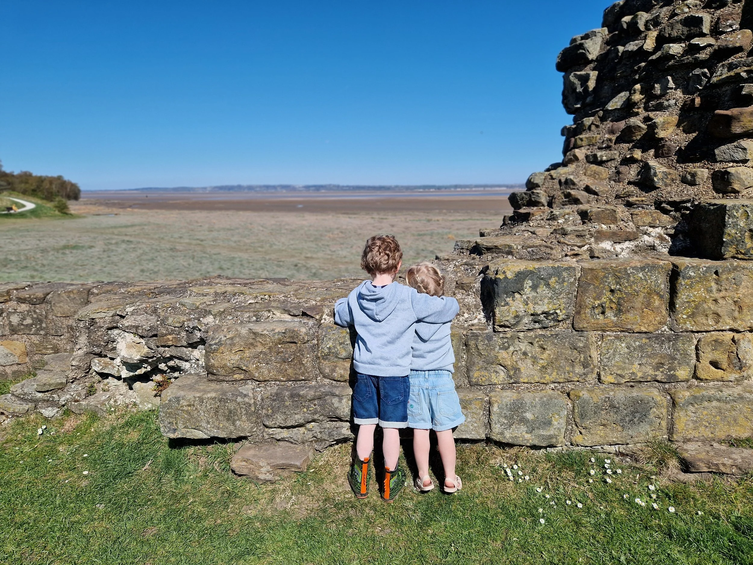 Two children, a boy and a girl, are standing side by side on a grassy area, facing away from the camera, looking at a large stone structure outdoors. The boy is wearing a gray hoodie and dark shorts, and the girl is wearing a light-colored dress.