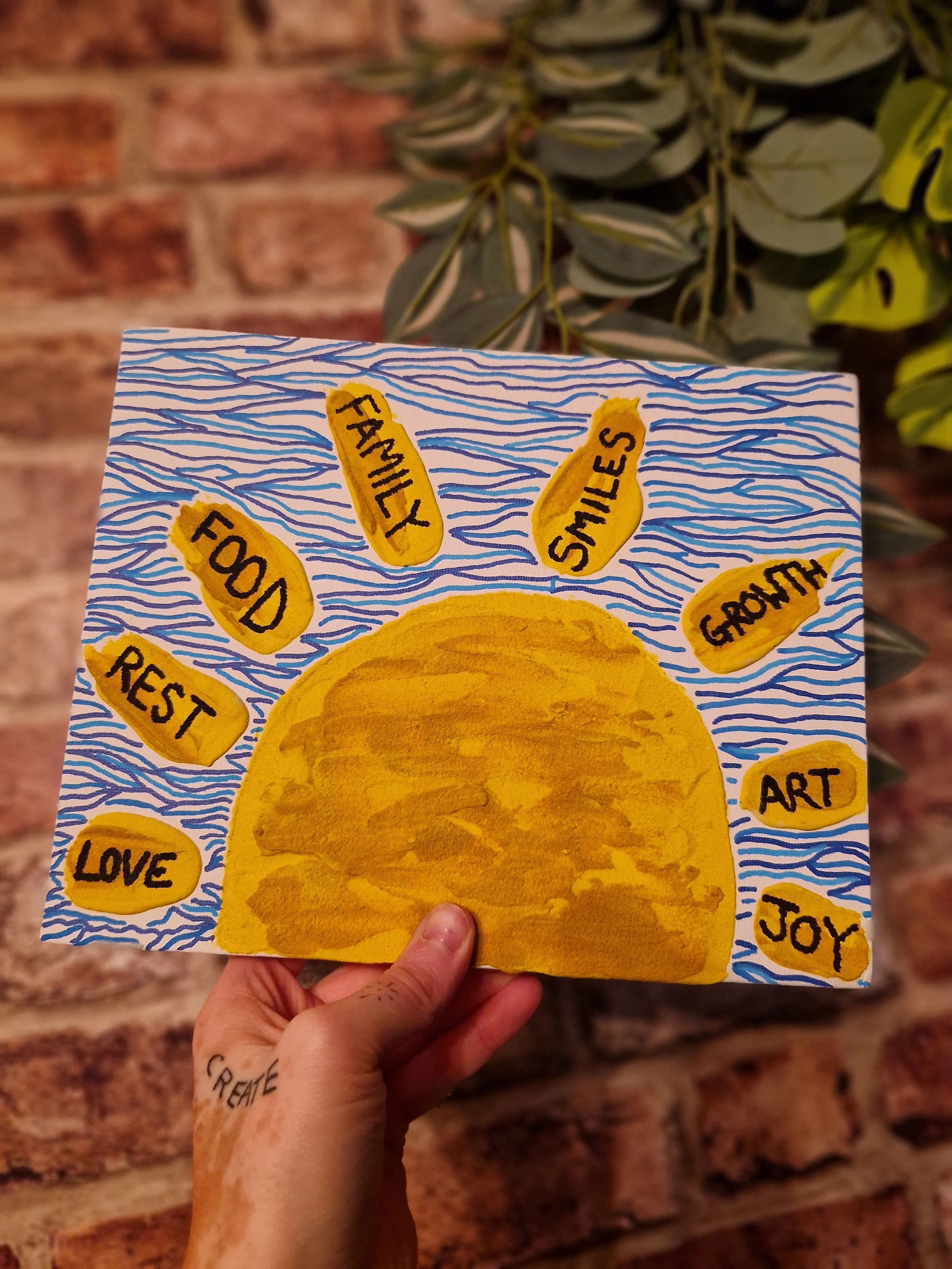 A hand holding a colorful postcard with a sun illustration and words like 'FOOD', 'FAMILY', 'SMILES', 'GROWTH', 'REST', 'LOVE', 'ART', 'JOY', and 'CREATE' written around it, with a background of a brick wall and green leafy plant.