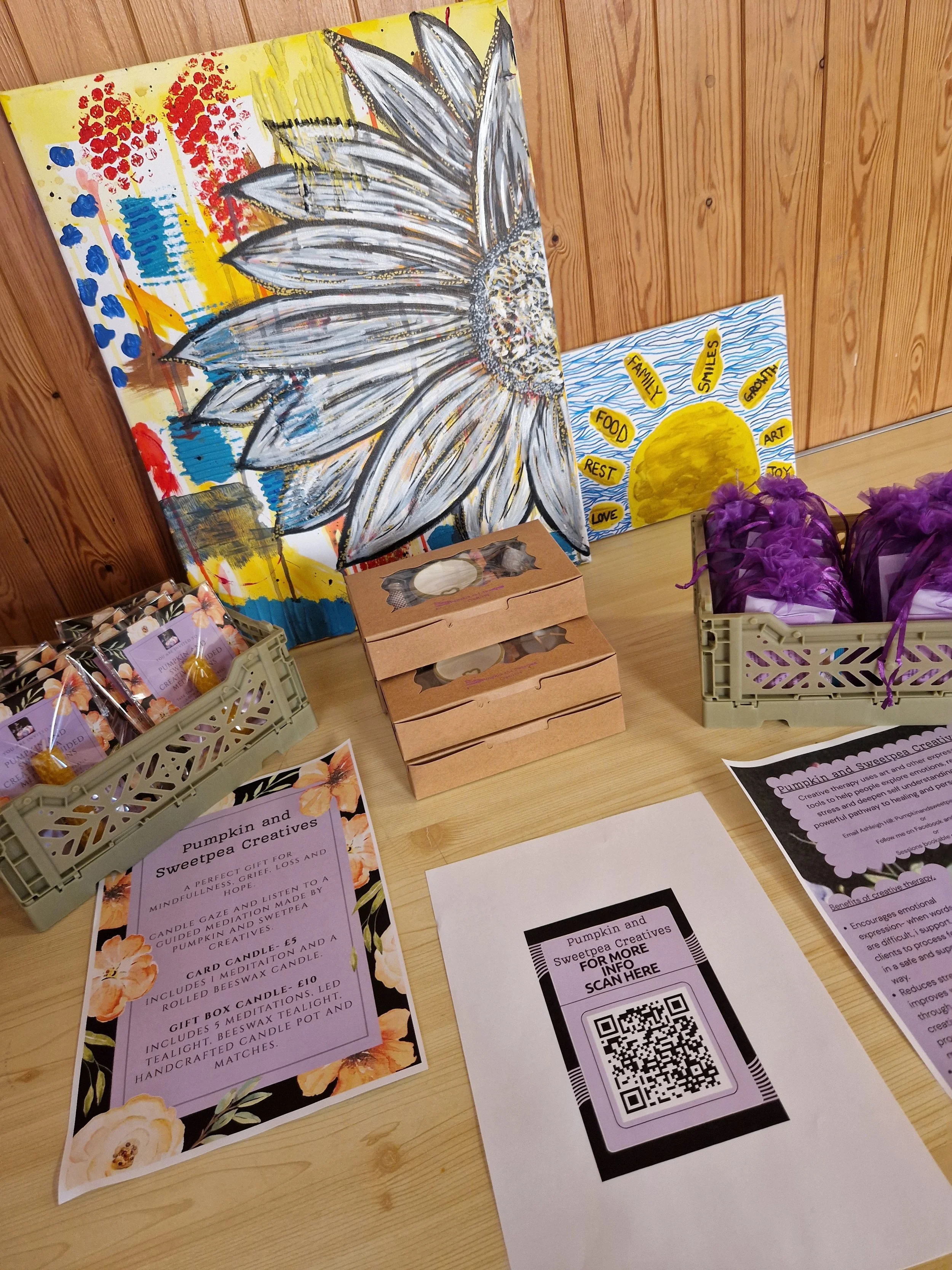 Display of promotional flyers and artwork for Pumpkin and Sweetpea Creatives, featuring a large painted sunflower, a colorful painting with red, yellow, and blue splashes, and a smaller poster with a yellow sun and handwritten words like 'family', 's
