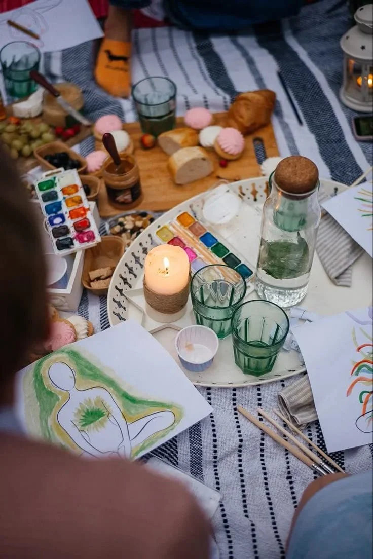 A cozy outdoor scene with a striped tablecloth, featuring art supplies like watercolor paints, paintbrushes, and drawings, along with snacks, drinks, candles, and a glass water bottle, suggesting a casual art and snack gathering.