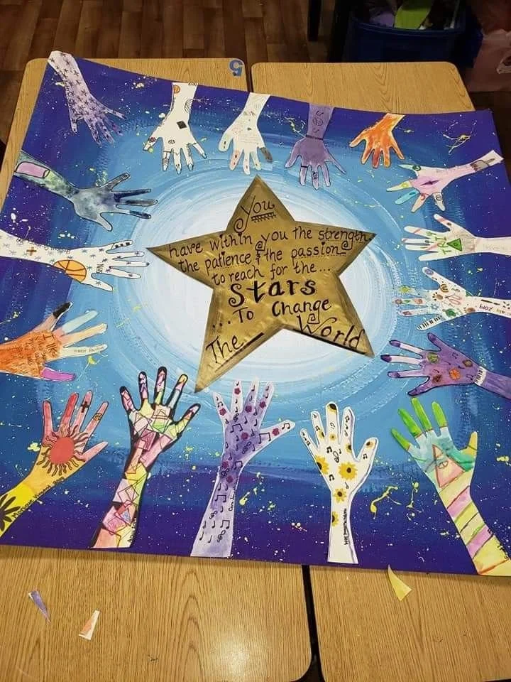 Colorful artwork featuring multiple handprints in various colors surrounding a golden star in the center. The star has an inspirational quote about strength and reaching for the stars, set against a blue background with white swirl patterns and small splashes of color.