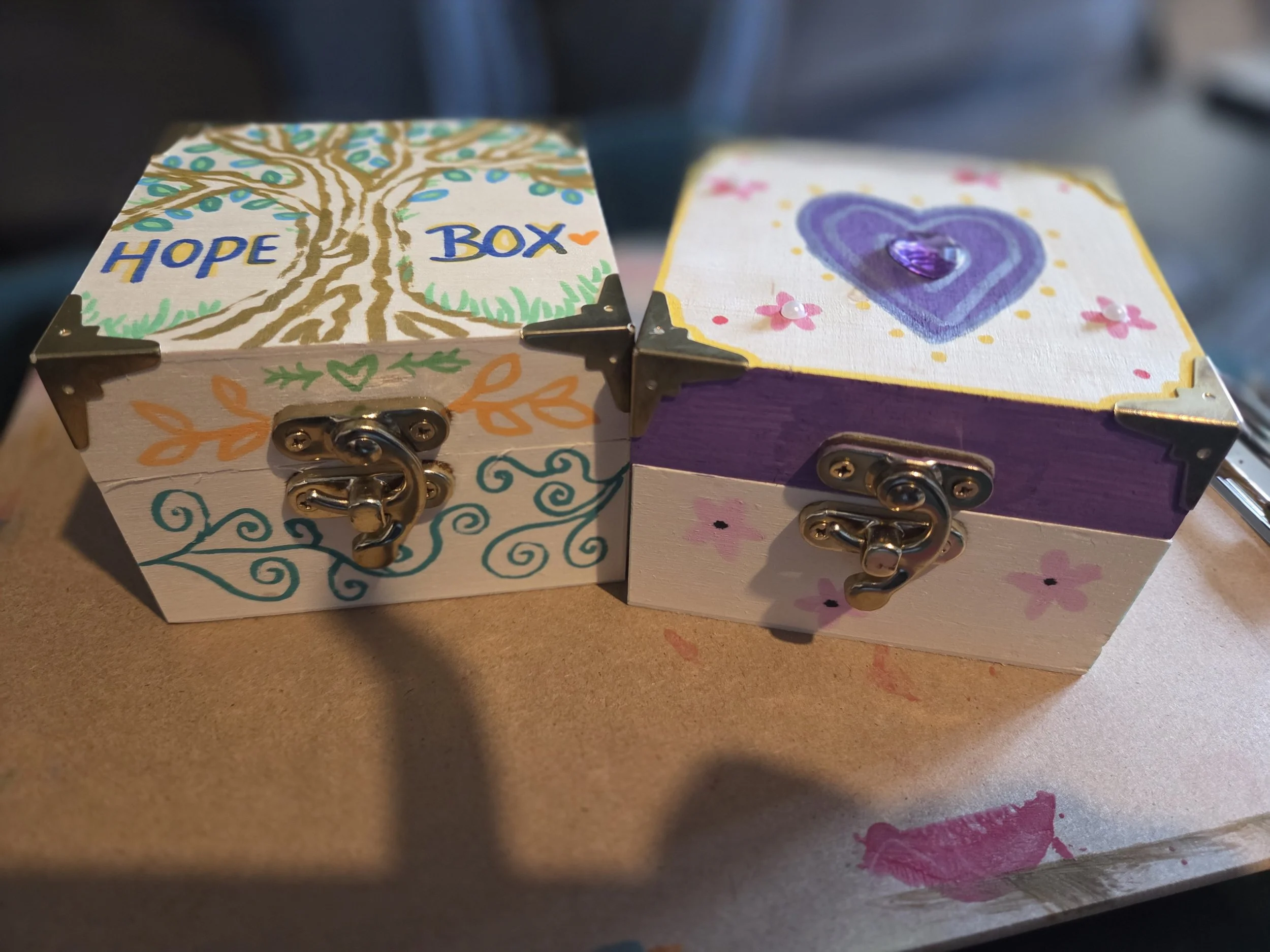 Decorative painted box with a tree and the words 'Hope' and 'Box' on the lid, and a separate painted cover with a purple heart outlined in black and small pink flowers, placed on a brown surface with pink and purple paint marks.