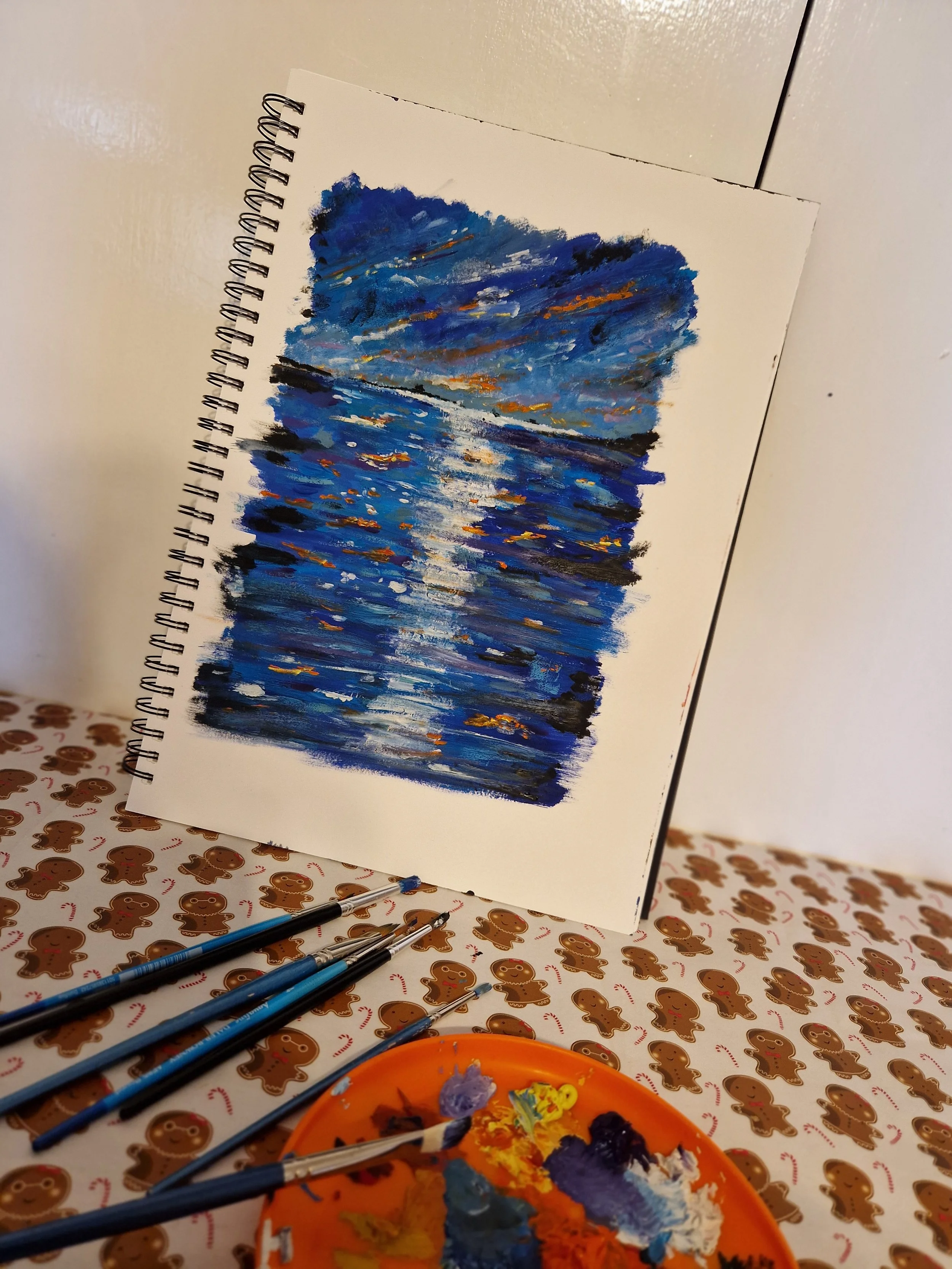 A painting of a sunset over a body of water on a spiral-bound sketchbook page, with painting supplies including brushes and a palette in the foreground, on a gingerbread-patterned table.