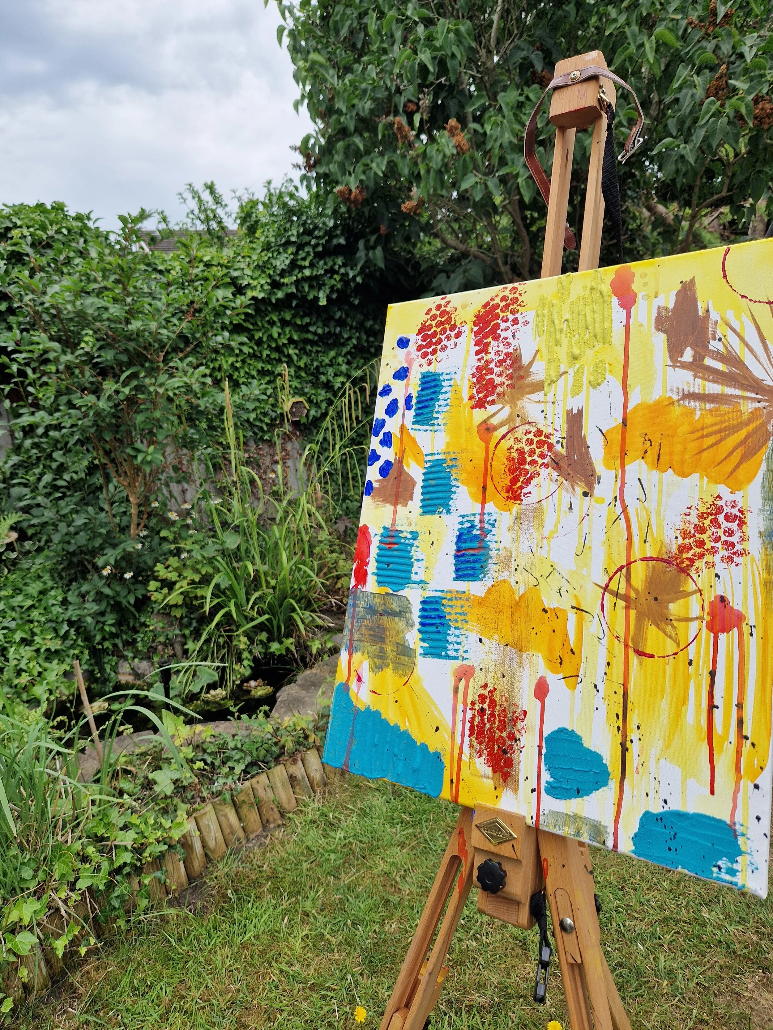 An outdoor scene with an abstract colorful painting on an easel, surrounded by greenery and a small pond with plants.
