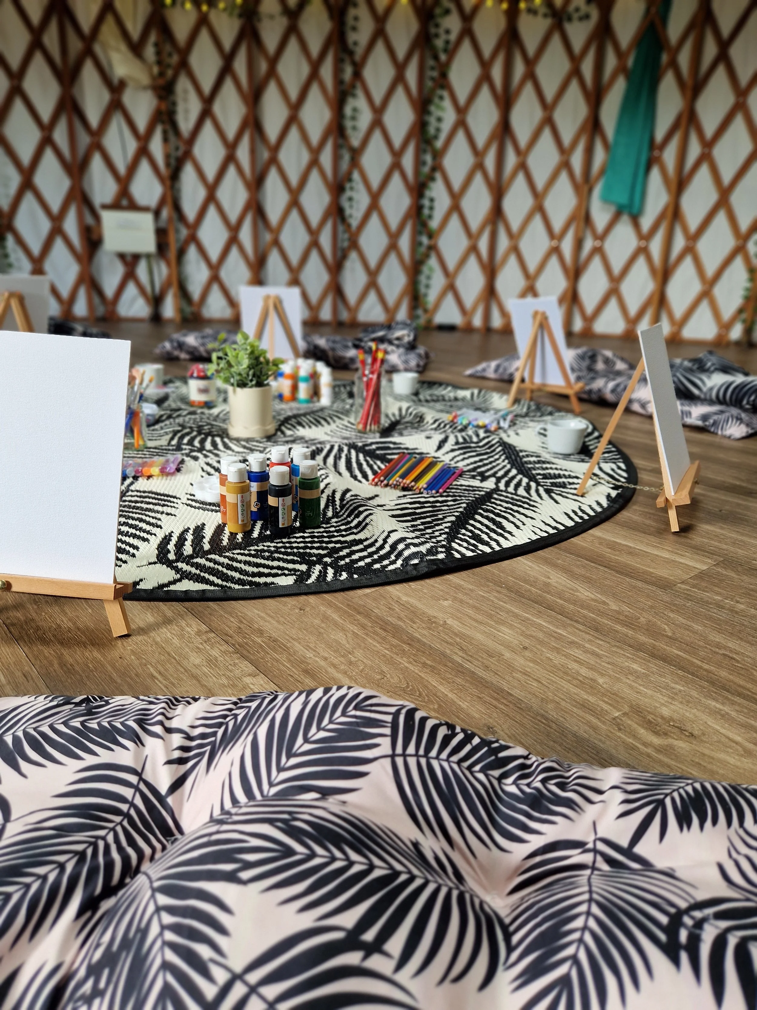 Workshop or art studio setup with a round zebra print rug, art supplies including paints, brushes, colored pencils, and small canvases, with pillows laid around and a lattice wall in the background