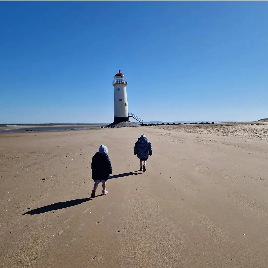 Two children walking on a sandy beach toward a lighthouse under a clear blue sky.