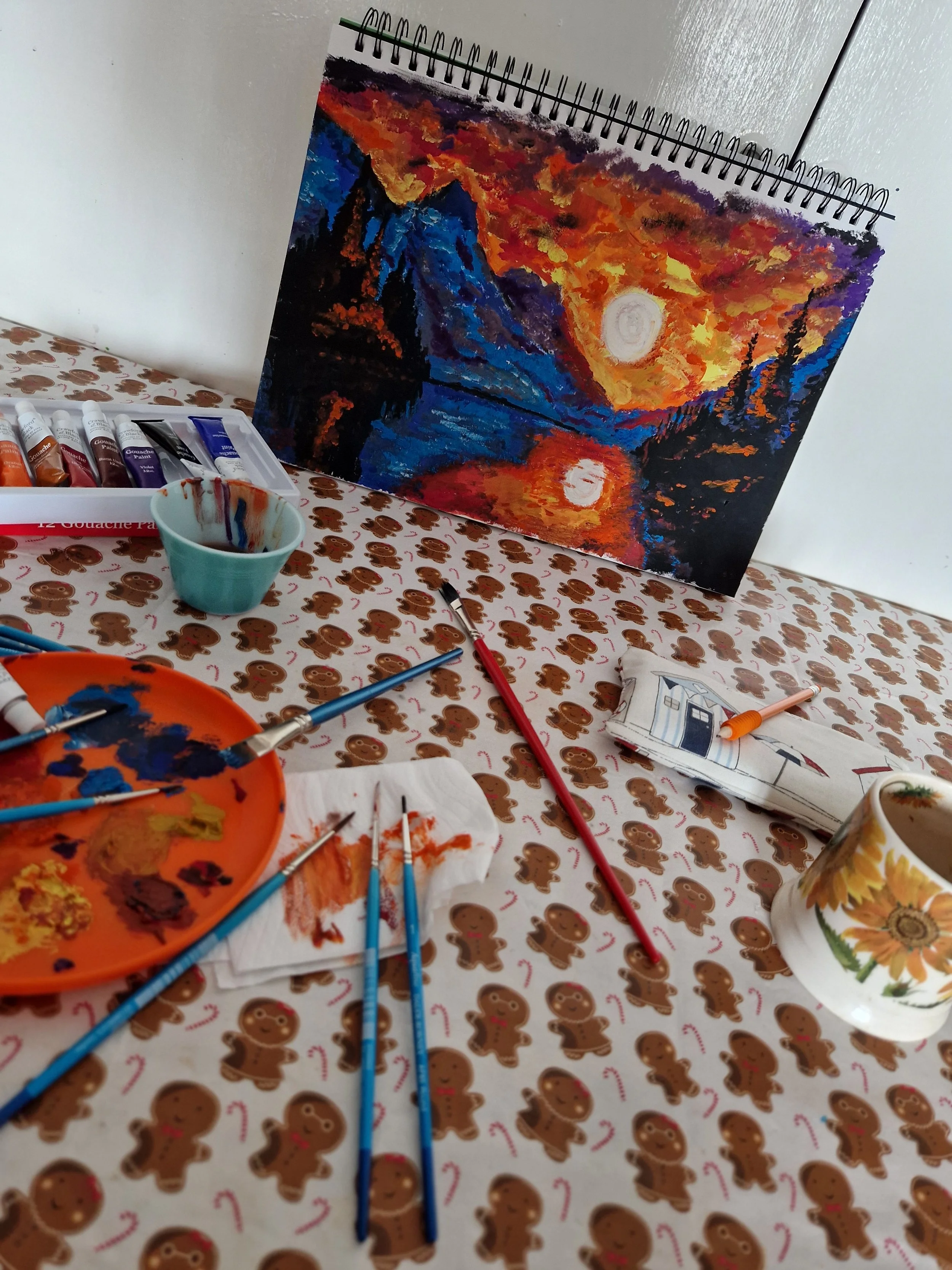 A colorful landscape painting of a sunset over mountains and a river. Painting supplies, including paintbrushes, paint tubes, a palette, and paper towels, are scattered on a table covered with a gingerbread men patterned tablecloth.