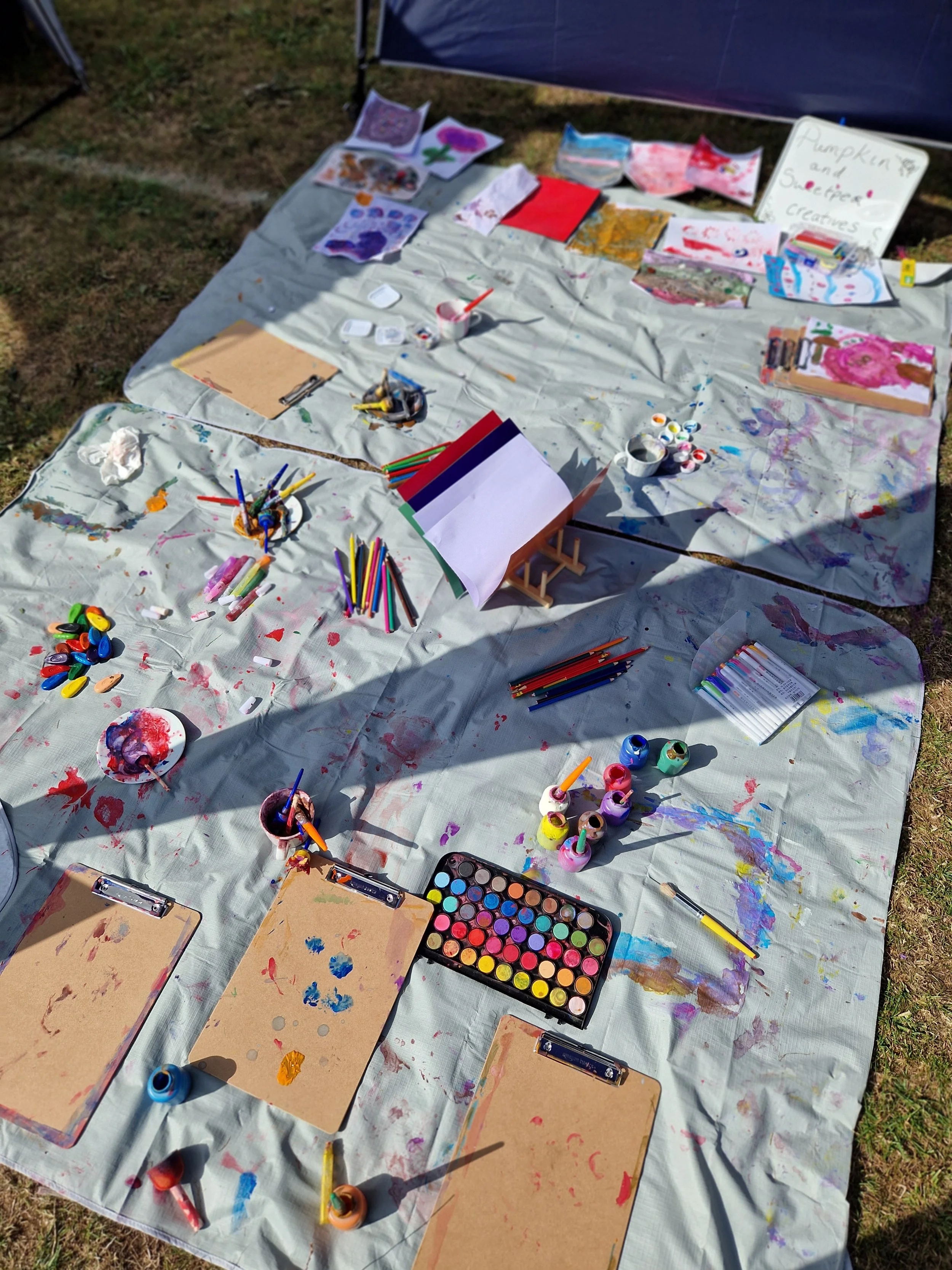Outdoor art setup with paint, brushes, markers, and paper on a large white tarp with grass around.