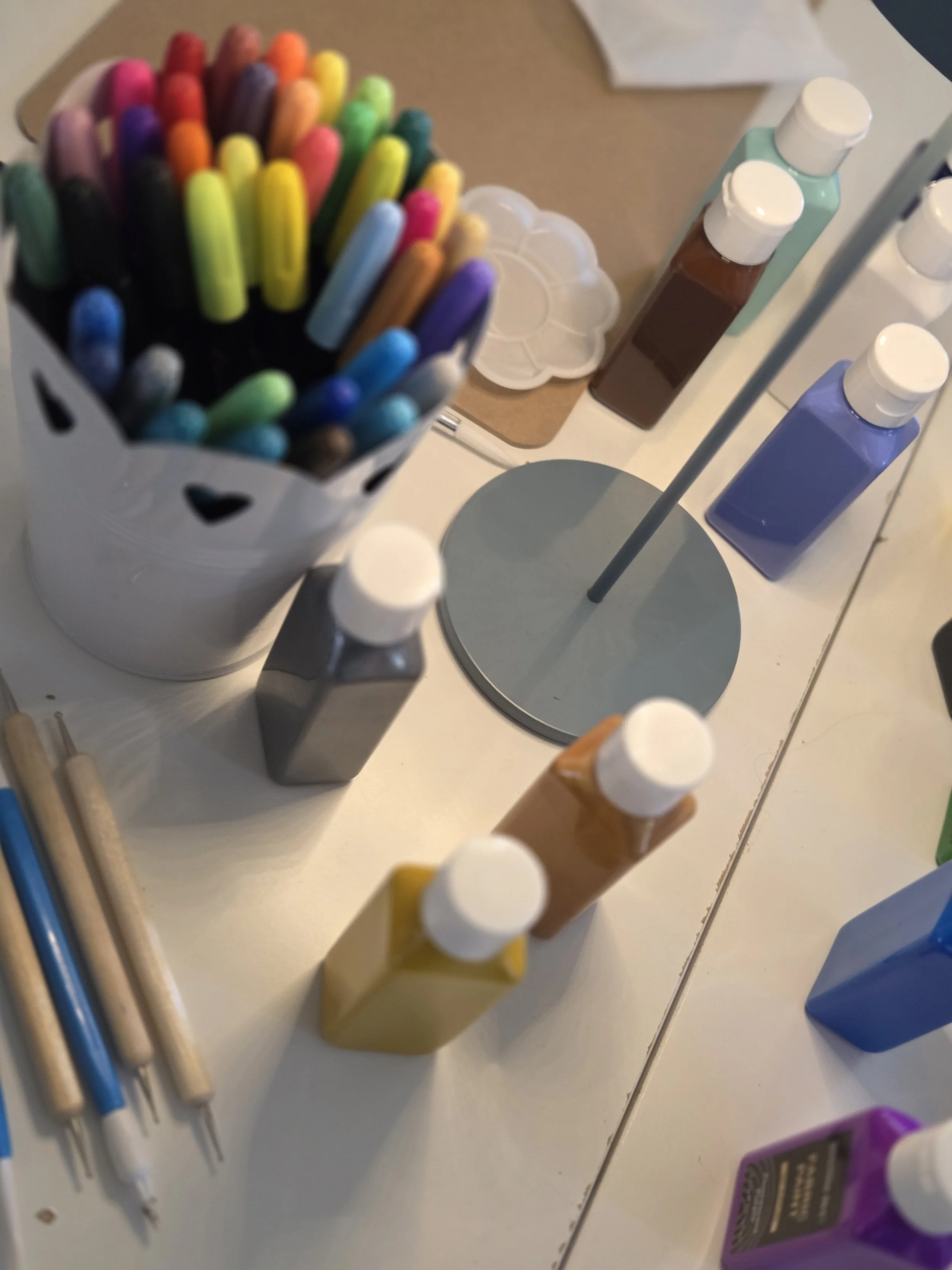 A collection of colorful markers in a white container, various bottles of paints, paintbrushes, and a color palette on a white surface.