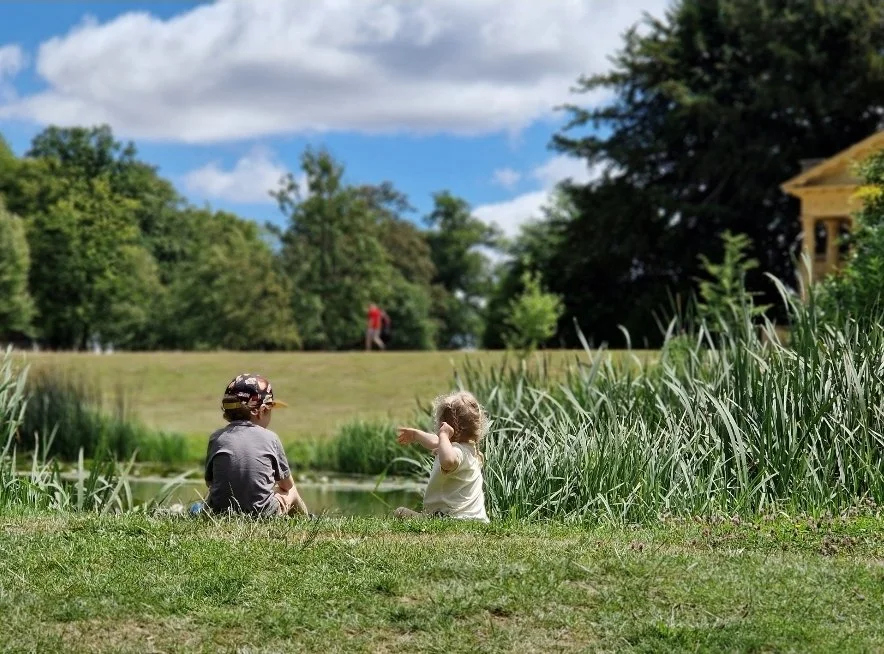 Two children, a boy and a girl, sitting near a pond, surrounded by grass and tall plants, with a park and trees in the background on a sunny day.