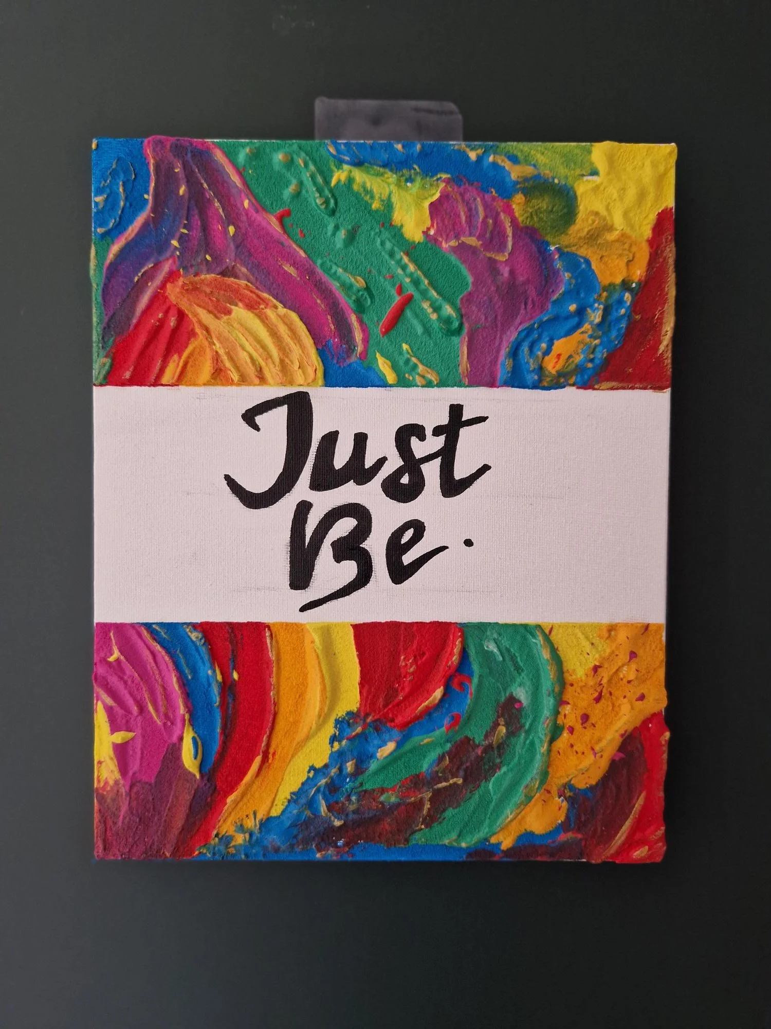 Colorful abstract painting with swirling patterns and splatters, with a white section in the middle containing the words 'Just Be' in black script.