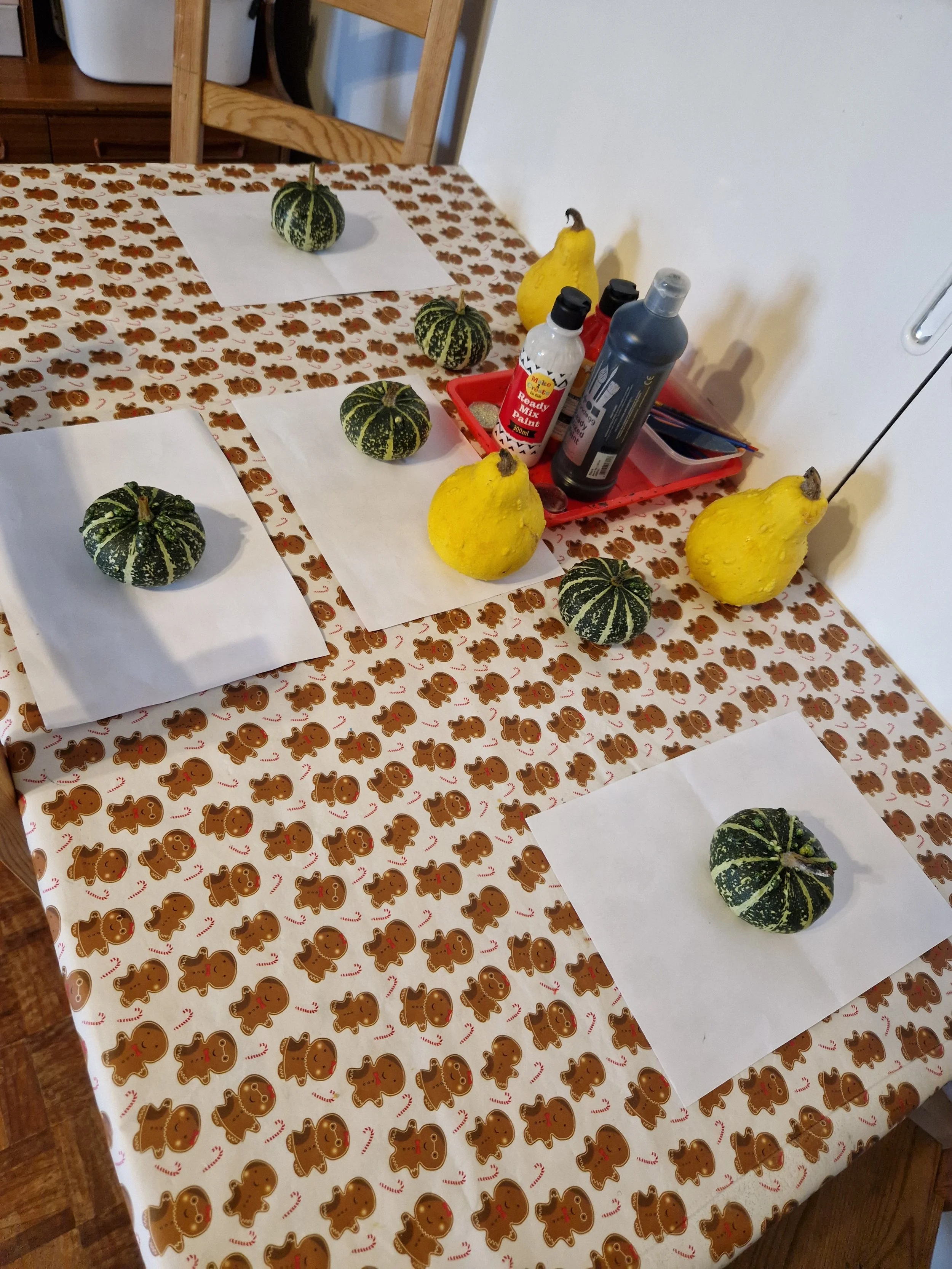 A table with a gingerbread men themed tablecloth featuring multiple small gingerbread cookies, each decorated with red and white details. On the table are white sheets of paper each topped with a small green and yellow striped pumpkin or gourd, and a