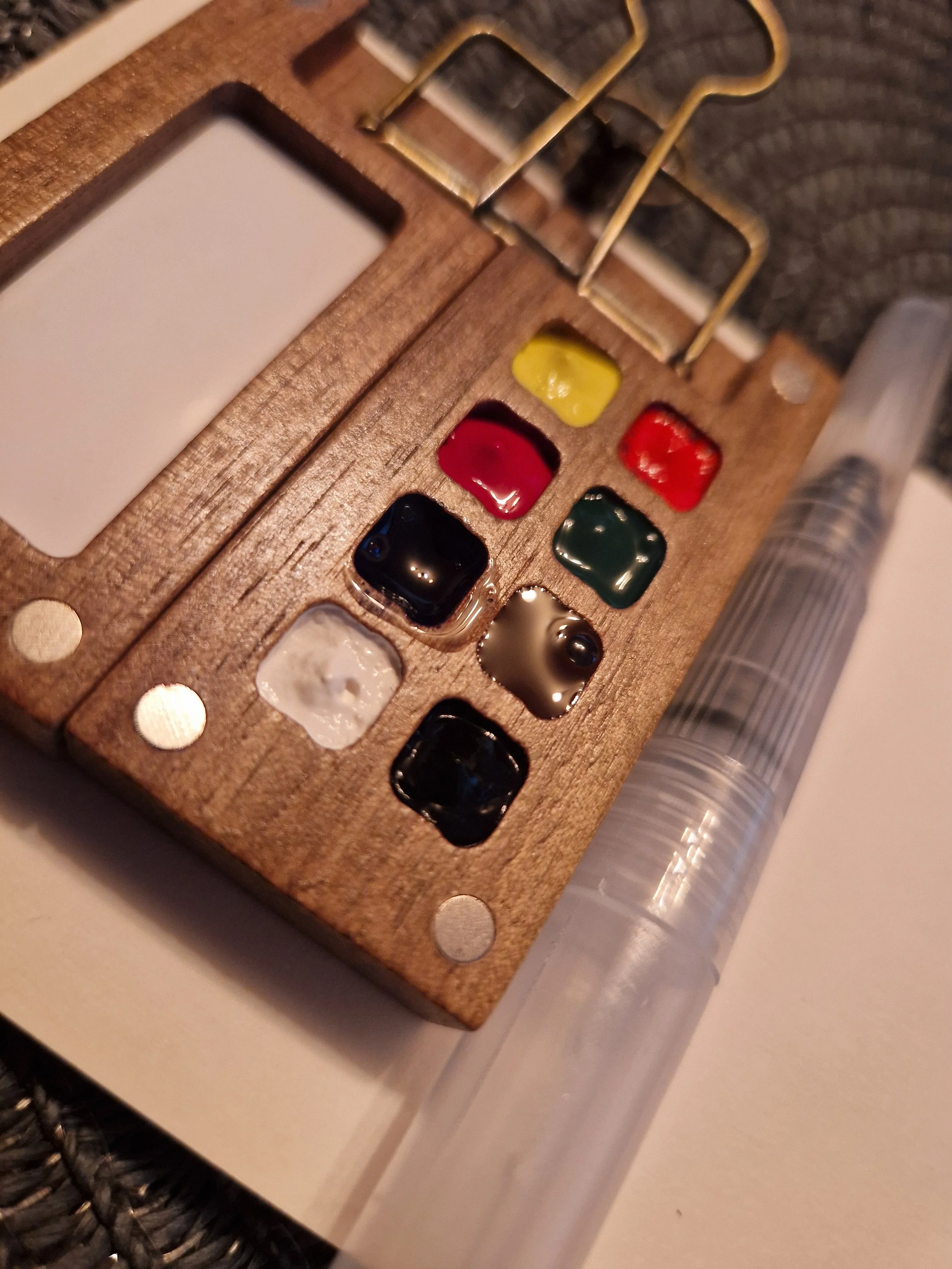 Close-up of a wooden palette with several small pools of different colored paints and a clear container of liquid on the side.