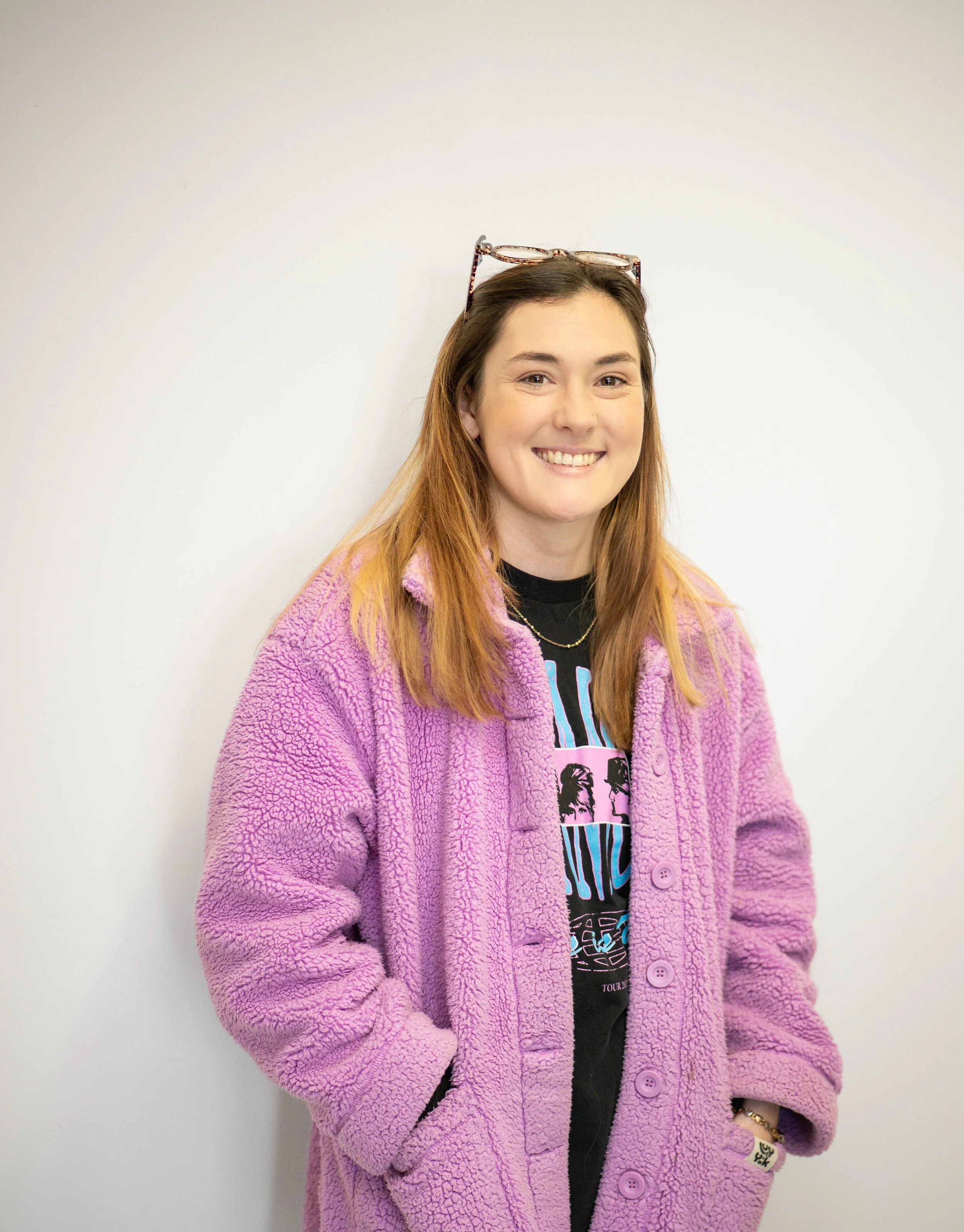 A woman with long, reddish-brown hair smiling, wearing glasses on her head, a pink fuzzy coat, a black graphic T-shirt, and gold jewelry, standing against a plain white wall.