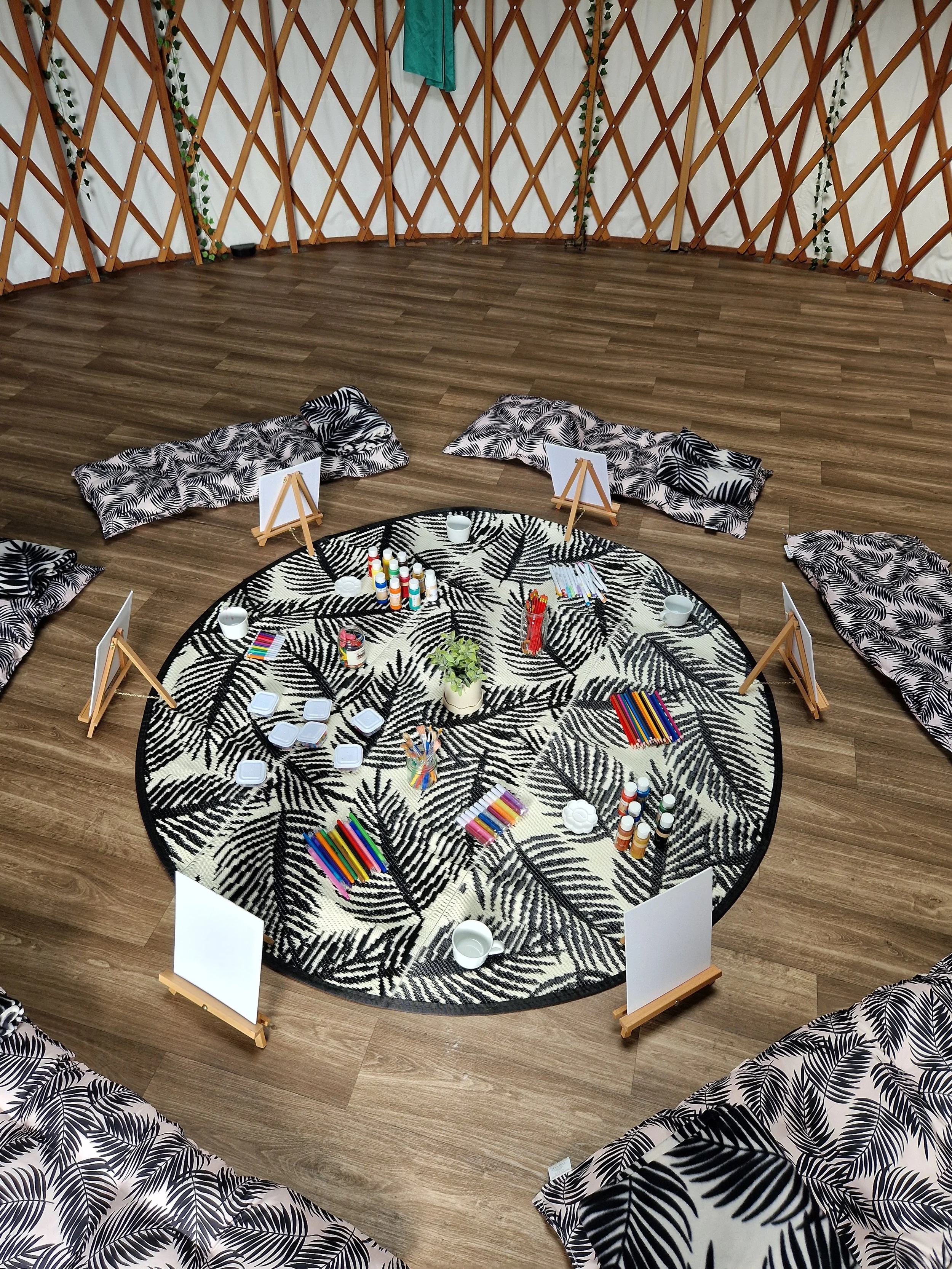 A decorated round indoor space with a black and white patterned rug at the center, surrounded by black and white leaf print cushions, small whiteboards on wooden stands, and various art supplies such as colored markers, paints, and palettes. The back