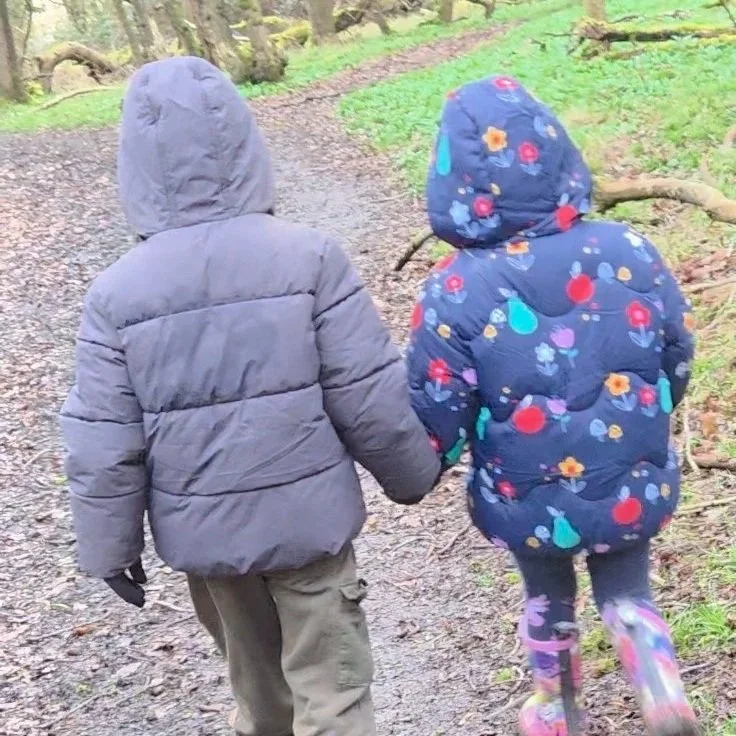 Two children, one in a gray puffer jacket and another in a dark floral jacket, holding hands and walking down a muddy forest trail.