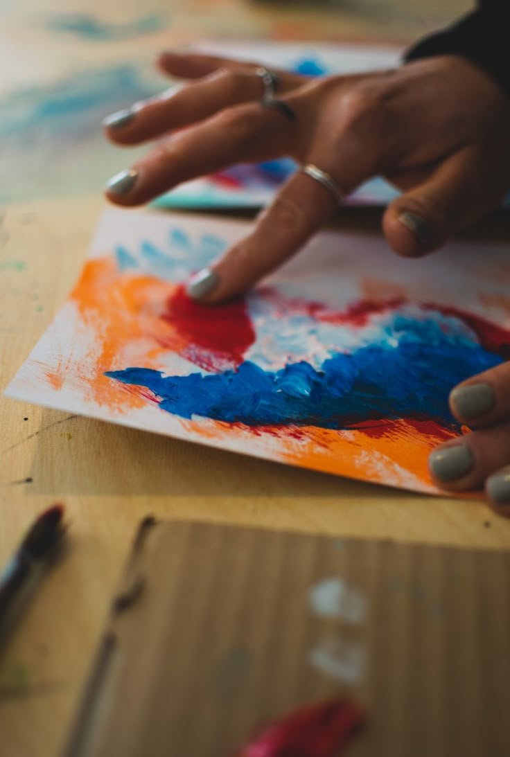 Close-up of a hand with silver rings, finger painted, creating colorful abstract art on a piece of paper with orange, blue, and red paint, on a wooden surface.