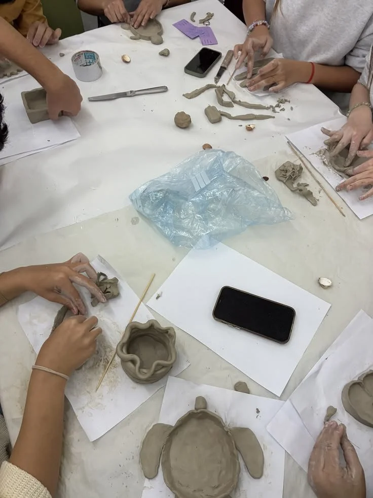 People working on clay pottery projects at a table, with various clay pots, tools, and phones visible.