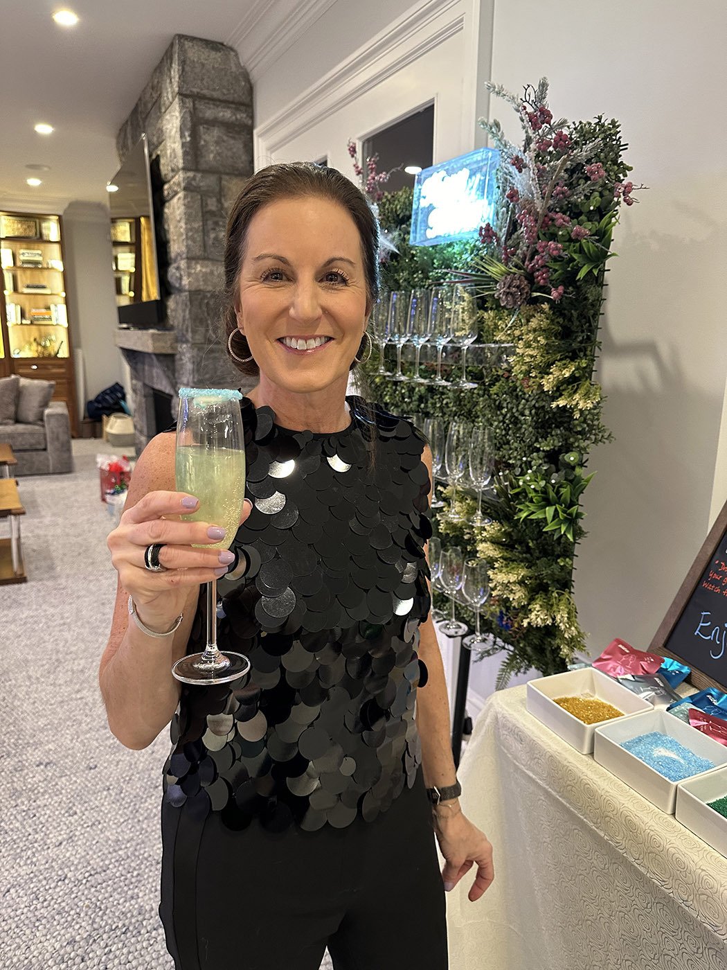 A woman smiling and holding a glass of champagne at a celebration event, with a decorated floral display and a sign in the background.