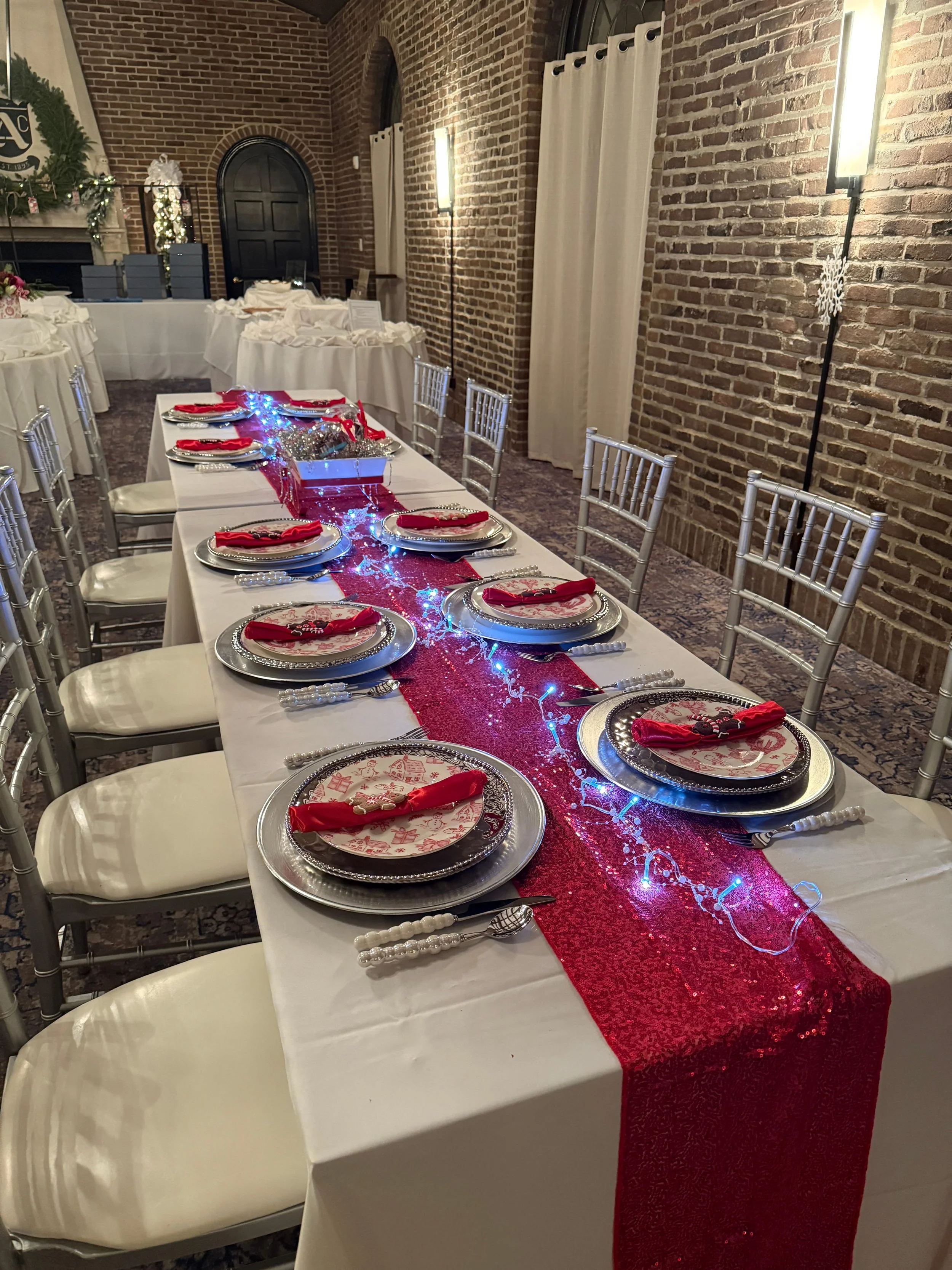 A banquet table set for a holiday dinner with a white tablecloth, red napkins, and a red sequined table runner decorated with fairy lights. The table has silver charger plates, patterned dinner plates, and silverware, with silver chairs arranged around it. The room has a brick wall, beige curtains, and warm lighting with modern wall lamps.