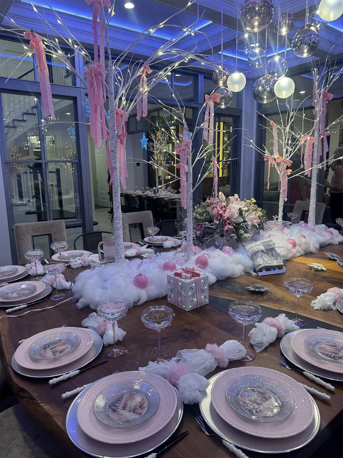 Elegant table decorated with white and pink accents, featuring a centerpiece of white trees with pink ribbons, hanging silver and white ornaments, surrounded by fluffy white material resembling clouds, with pink ornaments and wrapped presents on a wooden table, set for a celebration or event.