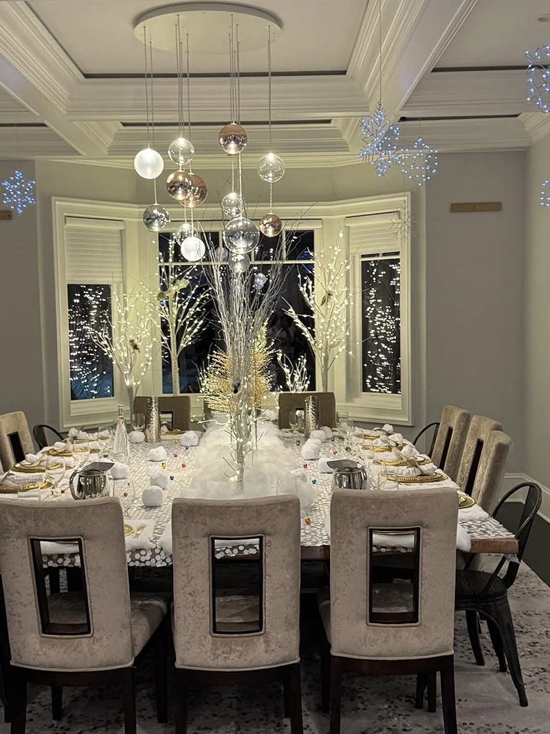 A festive holiday dining room setup with a large oval table decorated with a winter-themed centerpiece, white tablecloth, and gold accents. The room features snowflake decorations hanging from the ceiling, illuminated white tree branches outside the windows, and soft lighting creating a cozy, elegant atmosphere.