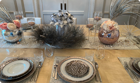 Elegant holiday table setting with silver and gold decorations, including a centerpiece of black feathers, a floral arrangement in a metallic vase, and decorative starburst sculptures, with plates, glasses, and cutlery set for a meal.