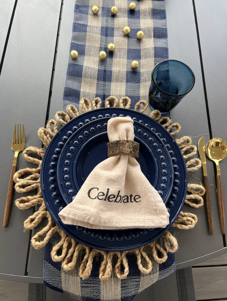 A table setting with a navy blue plate on a woven straw placemat, a beige cloth napkin with a bark ring, gold and black cutlery, a blue glass, and a blue and white checkered table runner with small round cheese balls.