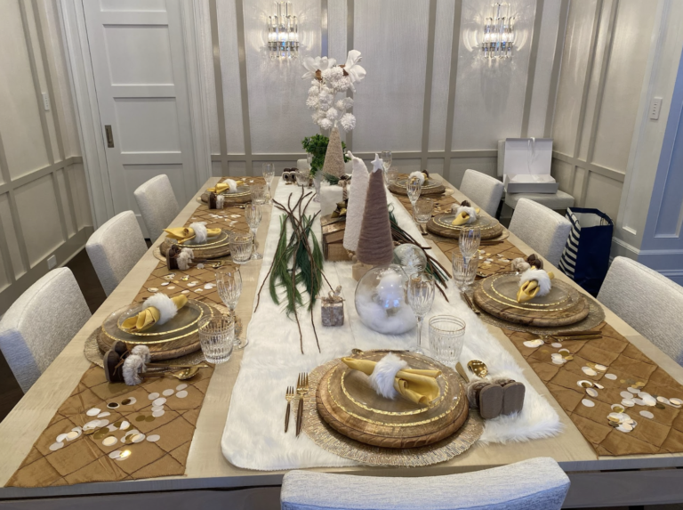 A decorated dining table set for a winter-themed celebration, with gold chargers, napkins with plush white sheep, clear glasses, and a centerpiece featuring white fluffy ornaments, tan and pink cone-shaped decorations, and eucalyptus branches, in a brightly lit room with white panel walls.