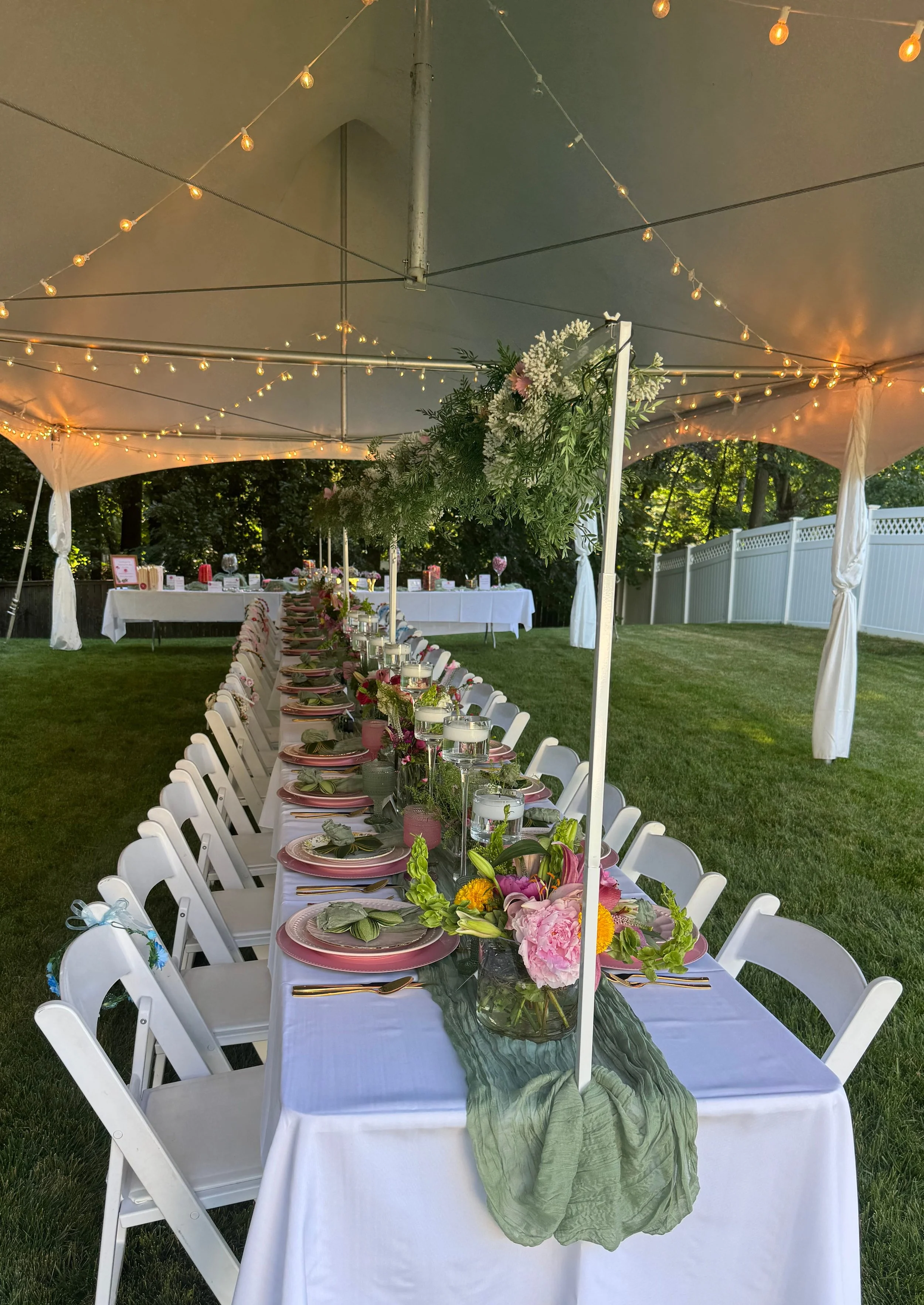 Long banquet table set outdoors under a large white canopy with string lights. The table is decorated with pink and green floral arrangements, pink plates, gold utensils, and glasses filled with water. A green fabric table runner is draped along the center of the table, and white chairs are arranged on both sides. In the background, additional tables with decorations are visible, as well as a white fence and trees.