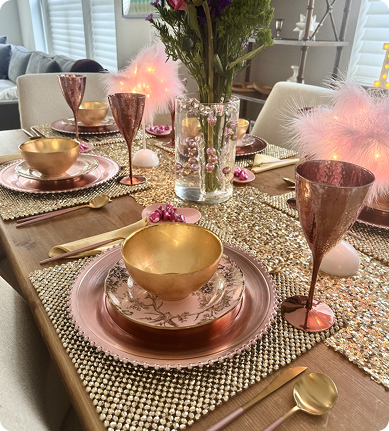Elegant dinner table setting with pink and gold dinnerware, copper goblets, gold utensils, a centerpiece of pink feathers, flowers, and decorative beads on a wooden table.