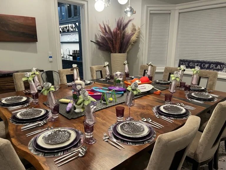 A decorated dining table set for a meal with black and white patterned plates, silverware, purple glasses, and cloth napkins with bows, in a room with a large vase of pampas grass, a sign that says 'The Gift of Play,' and windows with blinds.