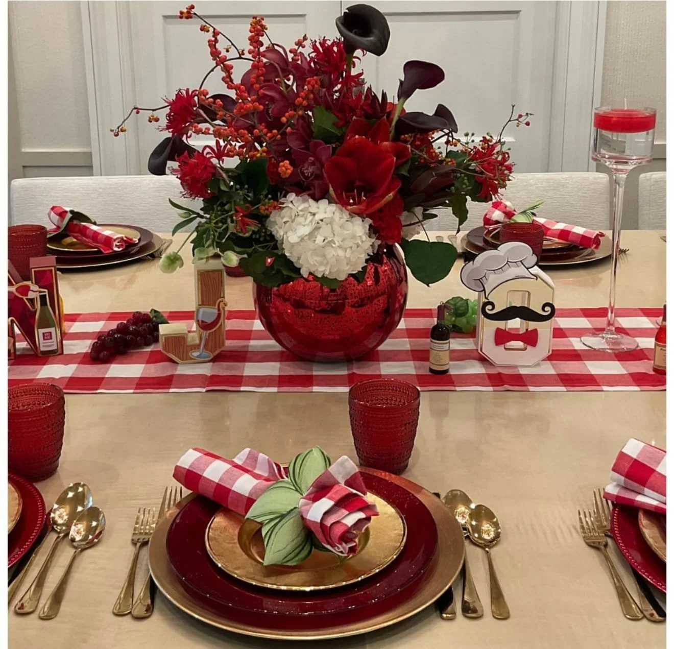 A dining table decorated for a festive occasion with a red and white theme. The centerpiece is a large red vase filled with red and white flowers. The table has red glasses, gold and red plates, red and white checkered napkins, and themed paper decorations, including a chef with a mustache. The table runner is red and white checkered, and there are small decorative items such as grapes, a small bottle, and a candle on the table.