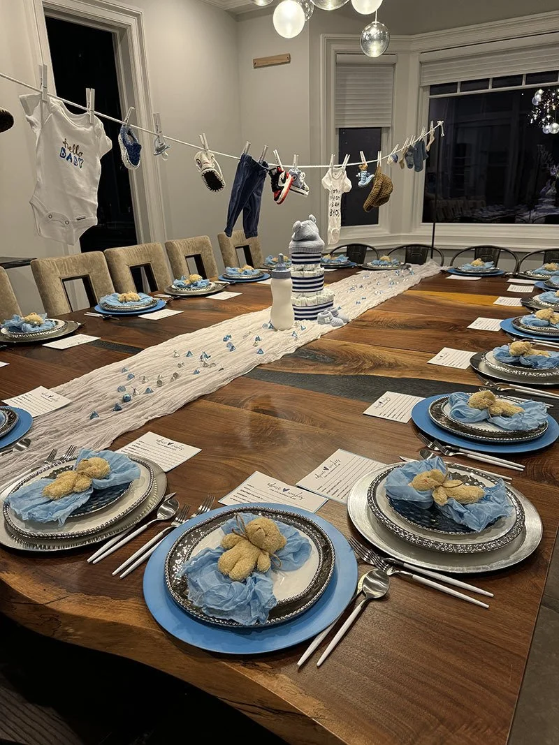 Baby shower table setting with blue and silver themed decorations, gift plates with teddy bear favors, a long white table runner with baby booties and tiny blue items, and a cloth line with baby clothes and socks hanging above the table.