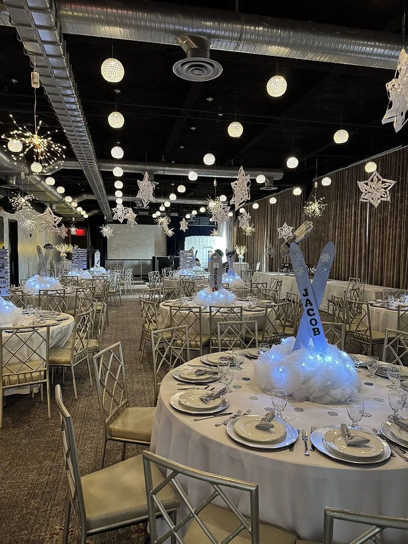 Event space decorated for winter or Christmas celebration with tables, white table settings, hanging snowflake decorations, and illuminated centerpieces resembling snow with oversized foam letters spelling 'JACOB'.
