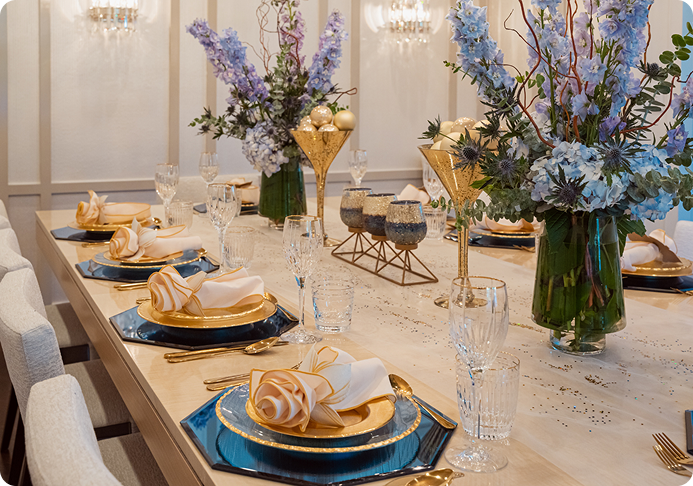 Elegant dining table set with gold-rimmed plates, ivory napkins folded with gold accents, crystal glasses, and tall floral centerpieces with blue and purple flowers.