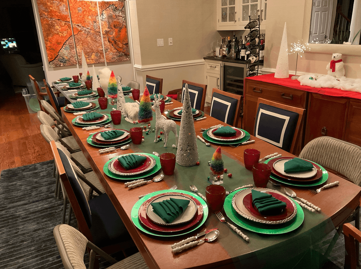 Christmas dinner table decorated with green and red plates, napkins, and red cups, with a holiday centerpiece of colorful felt trees, white reindeer, and snowball decorations, in a dining room with artwork and holiday decorations.