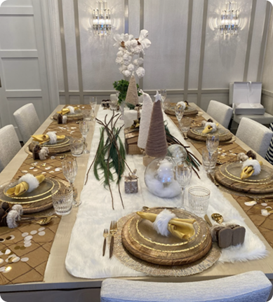 Elegant holiday table setup with gold and white decorations, including gold plates, napkins, and glassware, centered around a floral arrangement, with candles and festive ornaments in a decorated dining room.