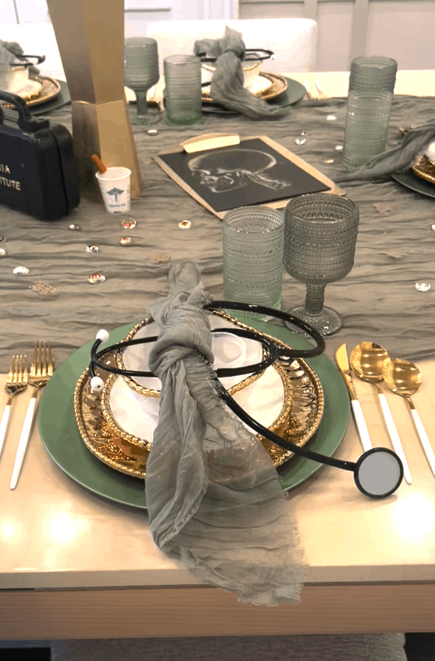 A hospital-themed table setting with gold and green dinnerware, a gray scarf on the plate, a stethoscope, and scattered rhinestones.