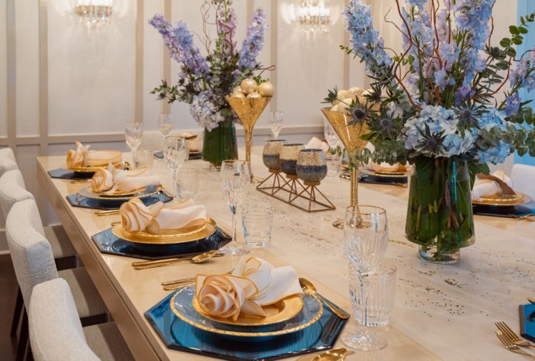Elegant dining table set with blue charger plates, gold-rimmed plates and napkins folded into roses, gold flatware, and tall crystal glasses. Centerpieces are large floral arrangements with purple and blue flowers in green vases, gold and glass candle holders, and decorative orbs. The background features white walls and wall sconces, creating a sophisticated atmosphere.