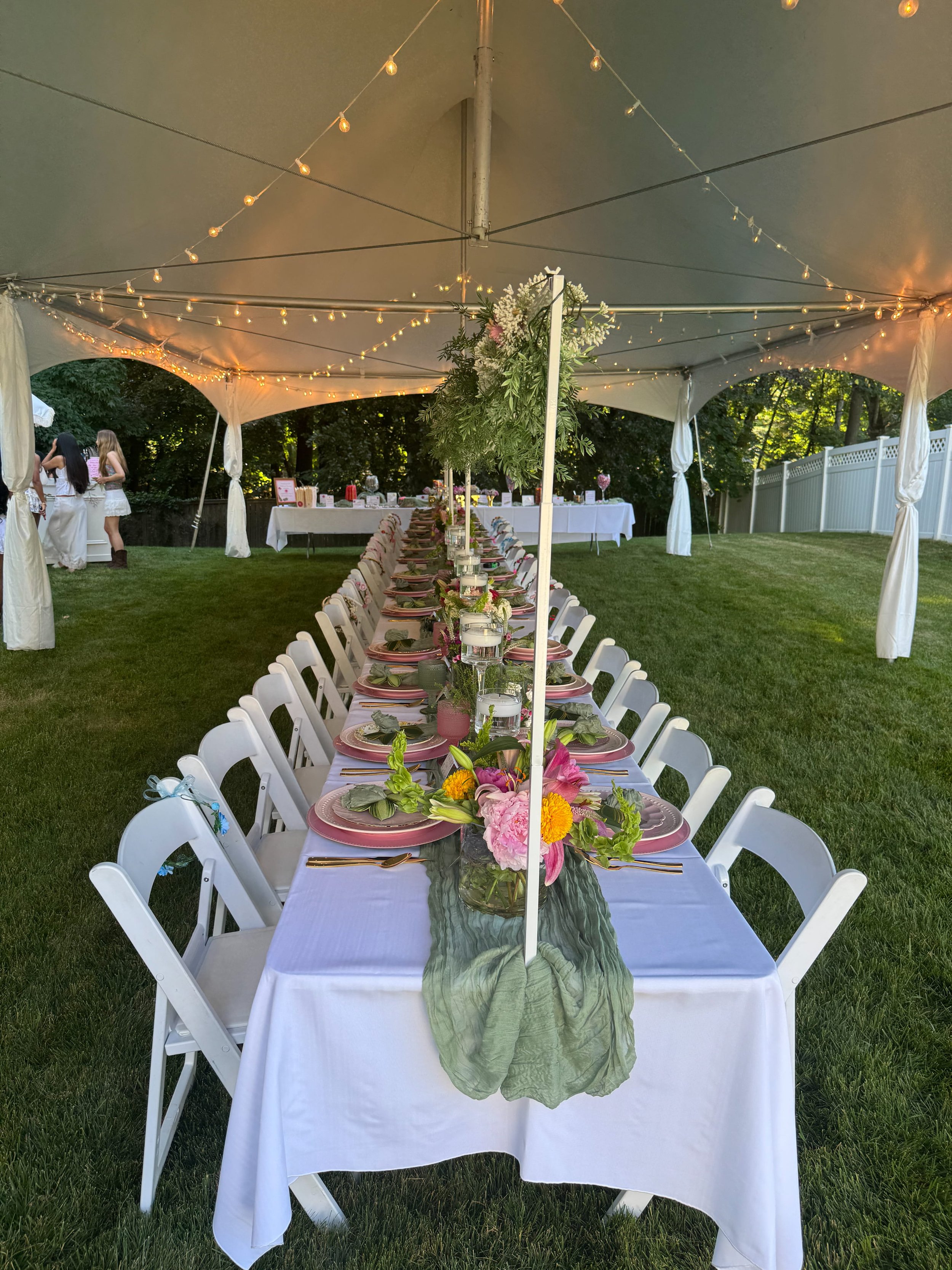 A long banquet table under a white canopy with string lights. The table is decorated with a green table runner, pink flowers, candles, and place settings. In the background, there are people standing and talking, with trees and a white fence surrounding the area.