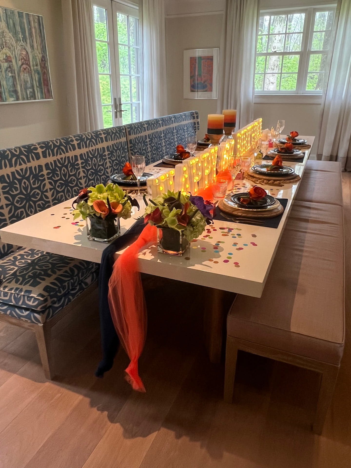 A dining table set for a celebration with black plates, orange napkins, and floral centerpieces, decorated with orange and multicolored confetti. There are three orange candles as a centerpiece, with a scalloped yellow light decoration on the table. Each place setting includes a wine glass and water glass. The table is surrounded by chairs, some with patterned cushions and others with plain cushions, near large windows with white curtains letting in natural light.