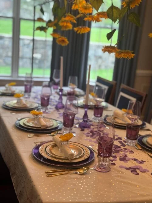 A decorated dining table set for a celebration, with purple themed glassware, plates, and purple flower petals scattered across a white tablecloth. Each place setting has a napkin and a small yellow flower, and there are tall candles on the table. The room has large windows with curtains, and hanging yellow flowers or decorations are visible above.