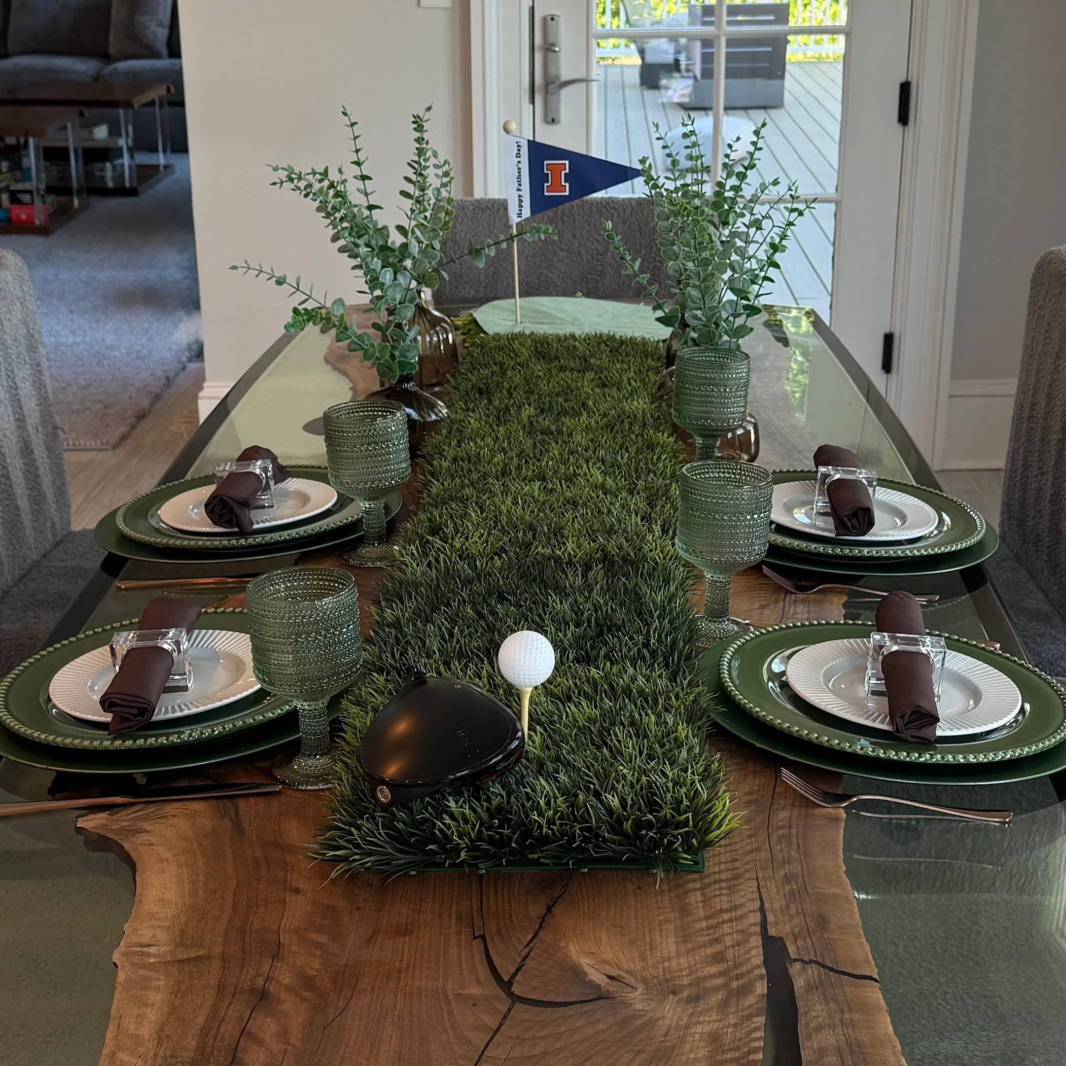A dining table decorated with faux grass, green candles in textured glass holders, and purple napkins, with a small plant and a miniature Iowa flag with the message 'Happy Y'dear! D. Day' on the table. The background shows a glass door leading to an outdoor deck.