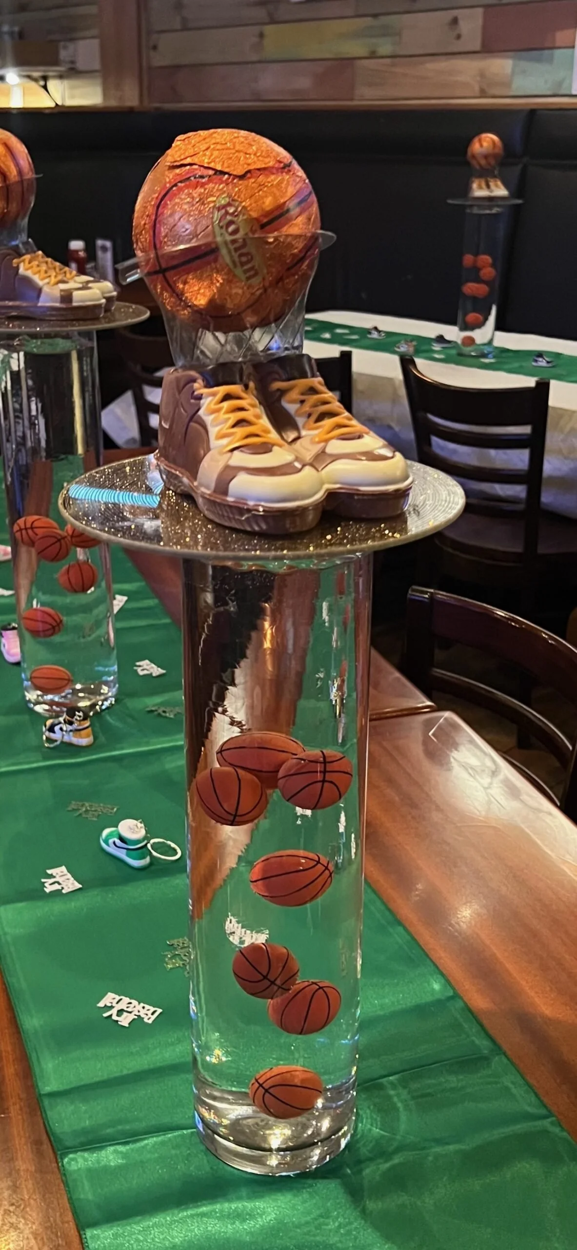 Decorative display featuring a glass of water with miniature basketballs inside, a pair of shoes with a basketball on top, and a basketball-themed table setting in a restaurant or party venue.